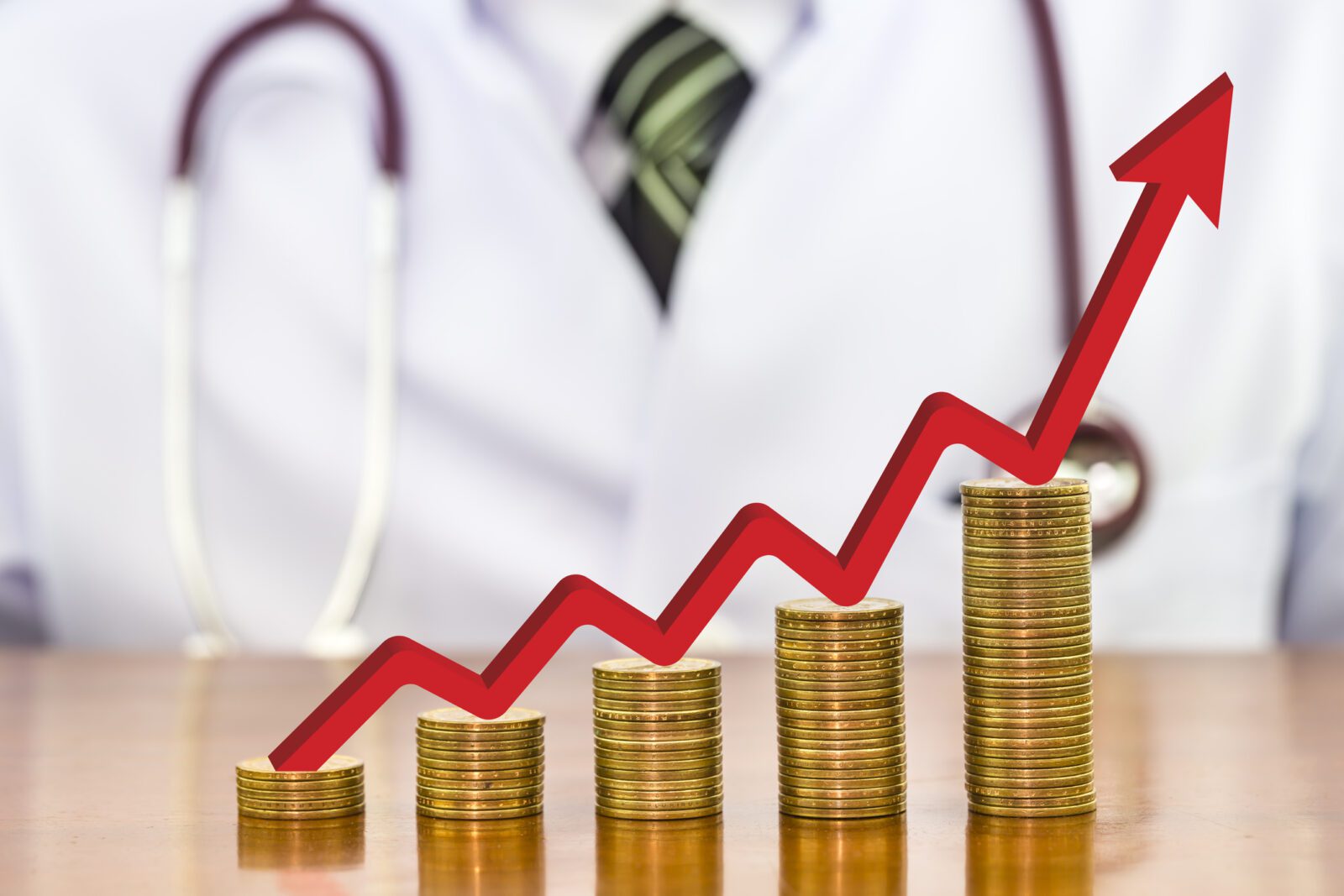 Red arrow over stack of money coins arranged as a graph on wood table with blurry the doctor stethoscope around neck a scene in the back, concept of financial health